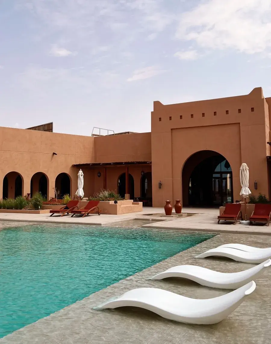 Resort pool with white in-water loungers in front of a terracotta building with arched gateways